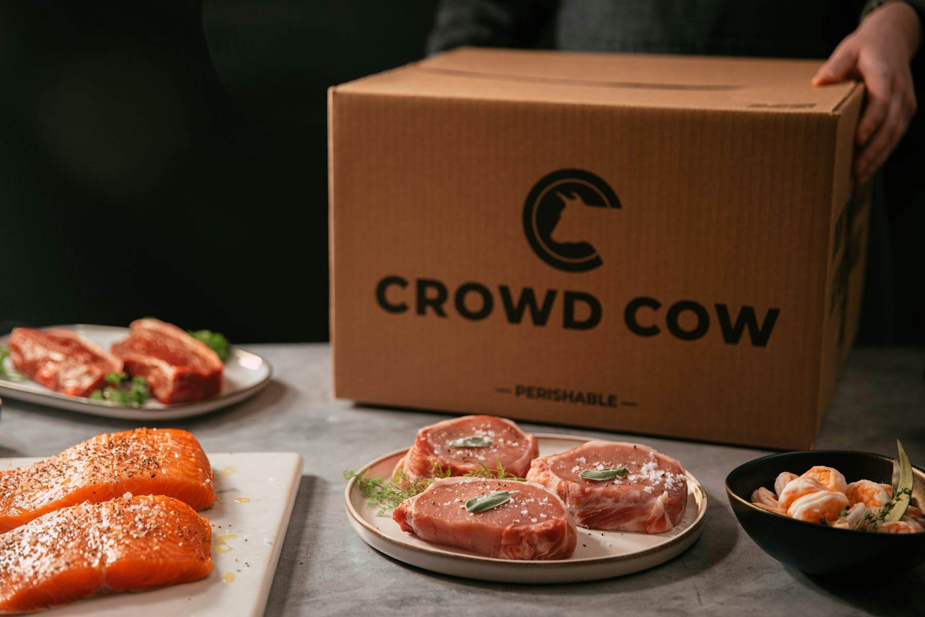 Photo of Crowd Cow meat -- porkchops, salmon, shrimp, and steaks -- in front of a gift box.