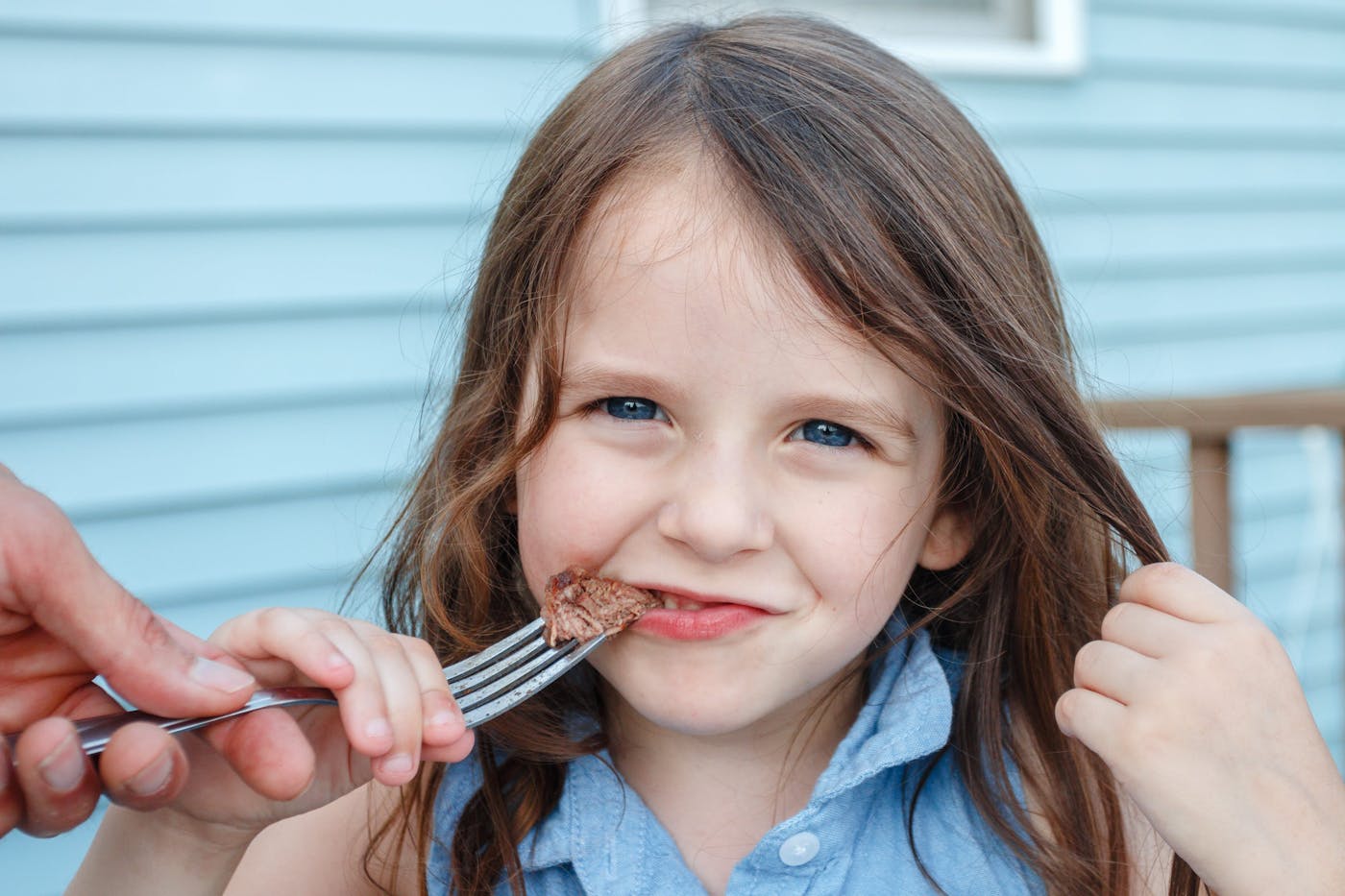 Child eating beef on a fork assisted by an adult.