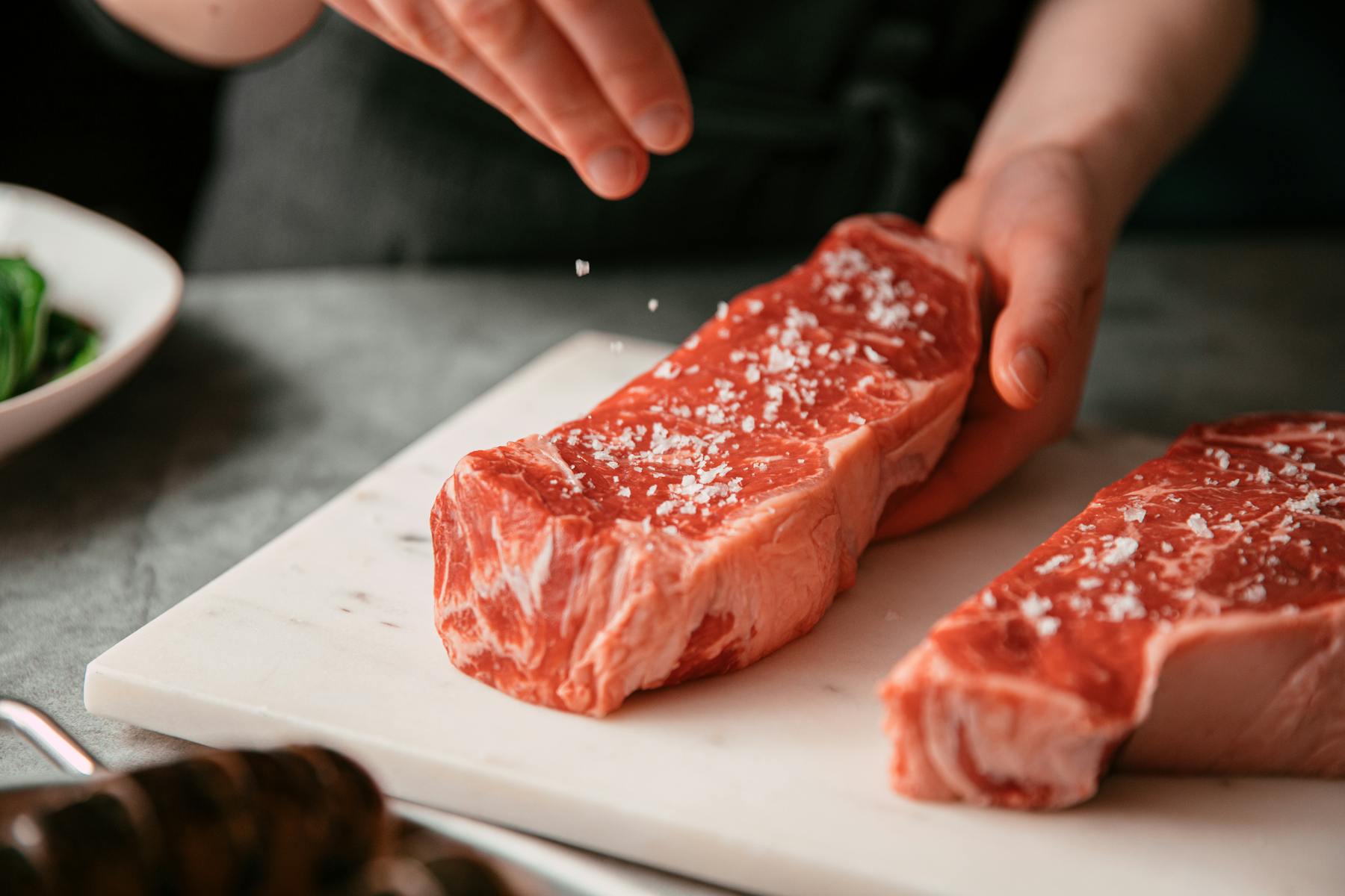 Image of two new york strip steaks being seasoned with salt.